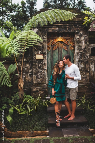 Fototapeta Happy couple in love smiling and kissing on background of traditional balinese architecture. Welcome to travel in Bali concept, vacations honeymoon in Asia.