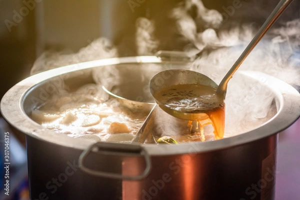 Fototapeta Cooking person in the restaurant is cooking while using the dipper in a large soup pot. And the water is boiling, there is smoke and hot spit.