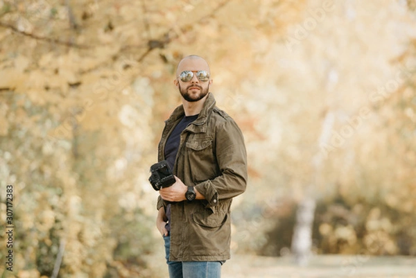 Fototapeta Bald photographer with a beard in aviator sunglasses with mirror lenses, olive cargo military jacket, blue shirt with digital wristwatch holds his camera and poses in the forest in the afternoon.