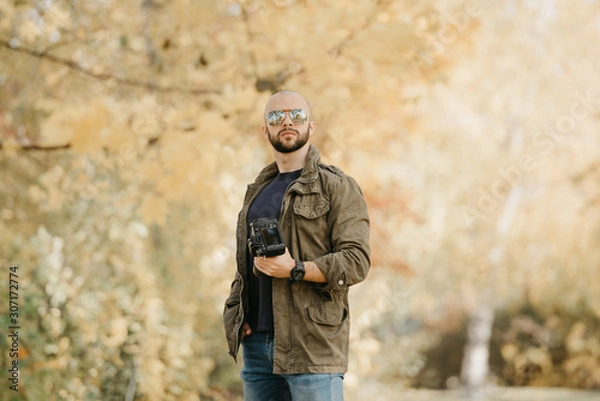 Fototapeta Bald photographer with a beard in aviator sunglasses with mirror lenses, olive military combat jacket, blue jeans and shirt with wristwatch poses holding his dslr camera and looks straight in the fore