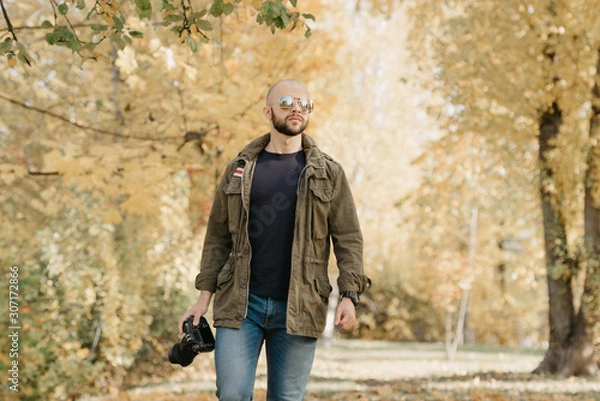 Obraz Bald photographer with a beard in aviator sunglasses with mirror lenses, olive military jacket, jeans and shirt with digital wristwatch holds the DSLR camera and goes straight on the battlefield 