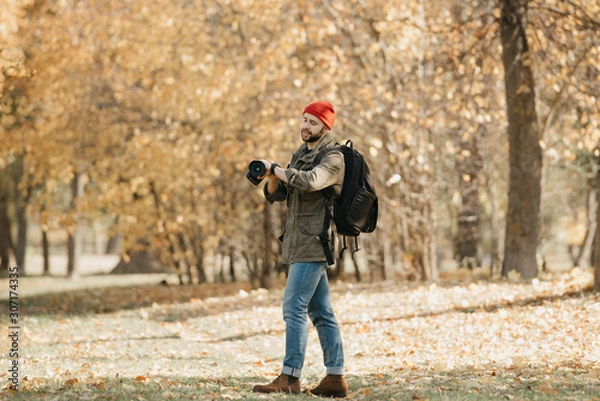 Fototapeta A photojournalist with a beard in an olive military cargo jacket, jeans, red hat with backpack holds his professional DSLR camera and checks the time on the wristwatch in the forest at the noon
