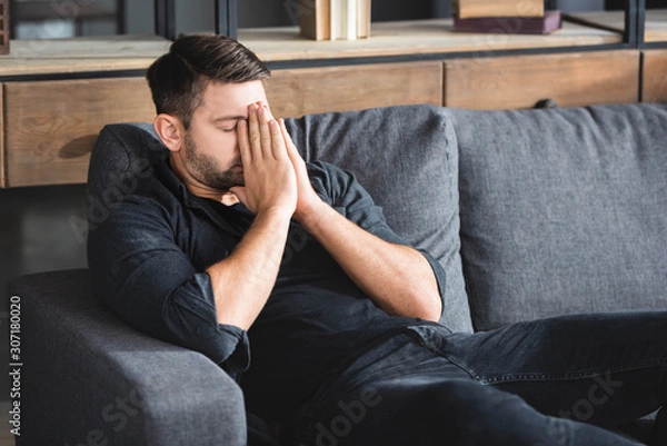 Fototapeta man with headache sitting on sofa and obscuring face in apartment