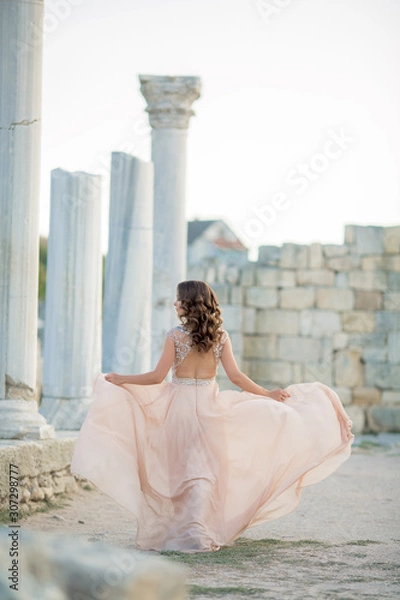 Obraz Adult woman between antique columns of architecture dressed in a long stylish light dress.
