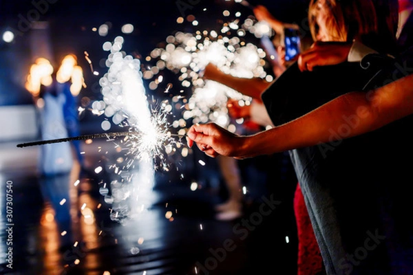 Fototapeta A crowd of young happy people with sparklers in their hands during celebration. Sparkler in hands on a wedding 2020 - bride, groom and guests holding lights in hand. Sparkling lights of bengal fires.