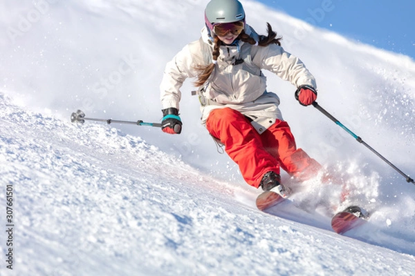 Obraz Girl On the Ski. a skier in a bright suit and outfit with long pigtails on her head rides on the track with swirls of fresh snow. Active winter holidays, skiing downhill in sunny day. Dynamic picture