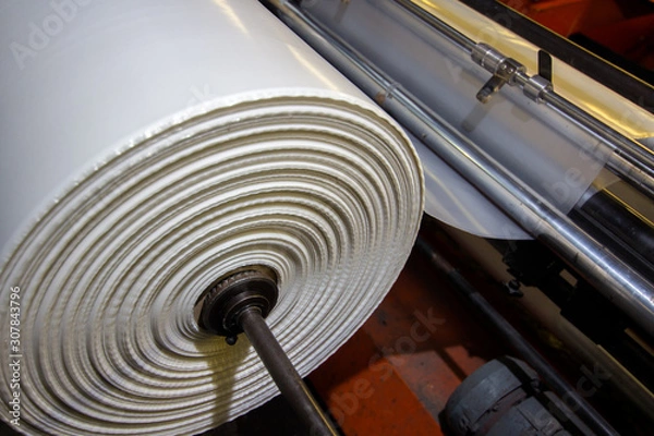 Obraz image of the inside workings of a machine with white high vis plastic sheet being fed through industrial factory on rollers with tension keeping it straight being stretched and cut ready for packing