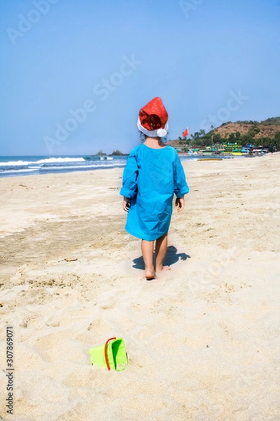 Obraz Little child in a Santa hat walks on a tropical beach.