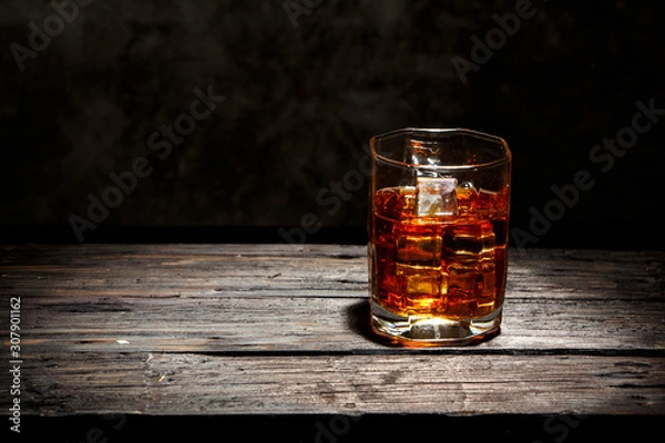 Fototapeta Whiskey with ice or brandy in a glass and a square carafe on an old wooden background. Whiskey with ice in a glass. Whiskey or cognac. Selective focus.