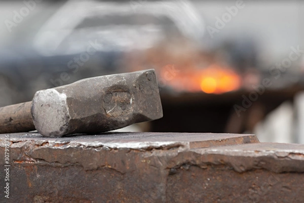 Fototapeta Old rusty hammer lies on the anvil with flame of brazier forge on background. Blacksmith, metalsmith, farrier tools.