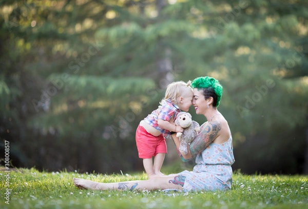 Fototapeta Young mother with green short hair is sitting on green grass, smiling, looking at her toddler son on her laps. Mother's day concept