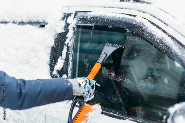 Obraz Close up photo of woman hand in grey mitten brushing the ice crust from the car window. Selective focus on scraper. Vehicle covered with snow. Heavy snowfall. Winter and seasonal concept.