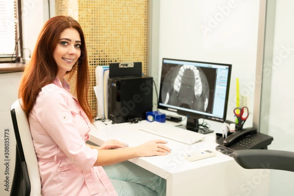 Fototapeta A female dentist doctor is sitting at a table, on a computer a CT scan of the jaw. The doctor is dressed in professional clothes.