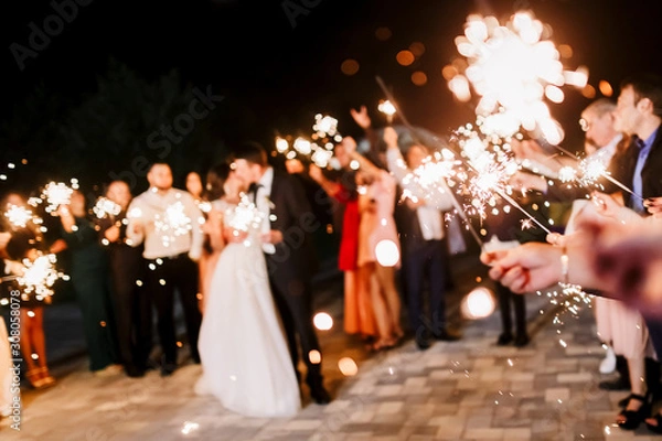 Fototapeta A crowd of young happy people with sparklers in their hands during celebration. Sparkler in hands on a wedding - bride, groom and guests holding lights in hand. Sparkling lights of bengal fires.