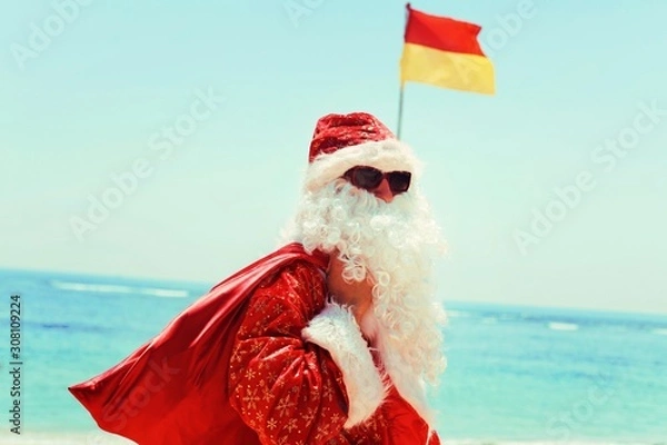 Obraz Santa Claus with a red bag of gifts near the Pacific ocean on a tropical island resting against the background of the Balinese flag