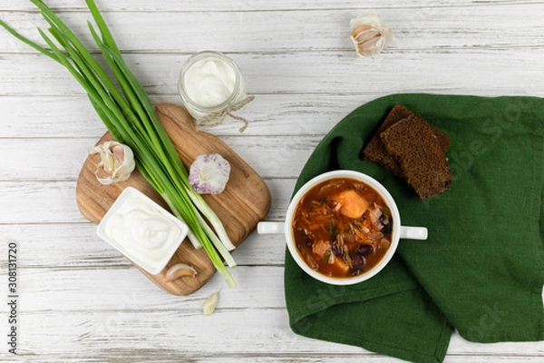 Fototapeta red borsch, a traditional national dish of Ukrainian cuisine. on the stock photo is served in a white pottery with cream, green onions and garlic on a light wooden background. green textile. flat lay