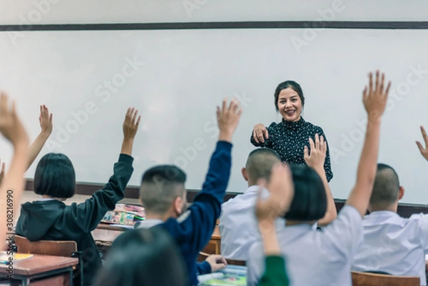Fototapeta A smiling Asian female high school teacher teaches the white uniform students in the classroom by asking questions and then the students raise their hands for answers.