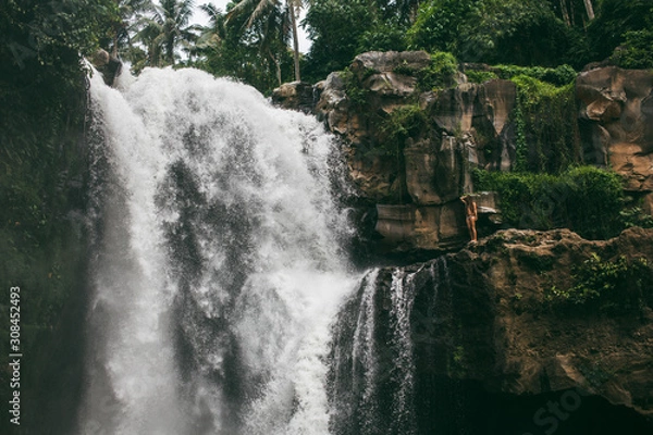 Fototapeta Beautiful girl posing in a swimsuit on the background of a large waterfall on the island of Bali. Tegenungan