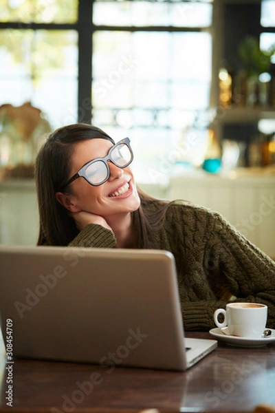 Fototapeta Stunning smiling positive caucasian brunette with glasses sitting in cafe and taking a break from work. On table are laptop and cup of coffee.