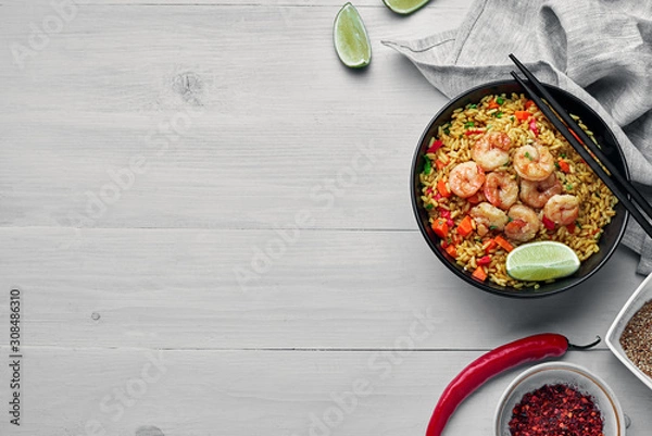 Obraz Delicious fried rice with shrimps, lime and vegetables in a black bowl. Traditional Asian food on a white wooden table with a fabric tablecloth. Top view, directly above shot with space for text.