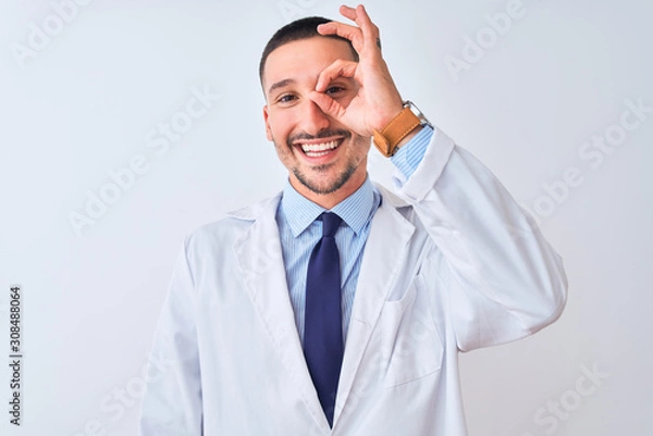 Fototapeta Young doctor man wearing white coat over isolated background doing ok gesture with hand smiling, eye looking through fingers with happy face.