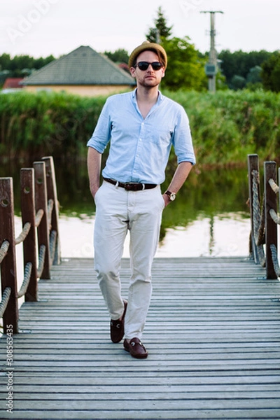 Fototapeta Handsome man in summer outfit wear sunglasses and hat posing near the lake in the city