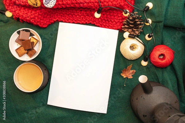 Obraz Christmas mockup, with a postcard, wishes, letter on the blanket with a kettle, leaf, tea, cone, chocolate and xmas tree lights.  Background top view blank space for text.