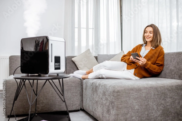 Fototapeta Young woman relaxing on the couch while air humidifier or purifier working on the foreground. Controlling it with a smartphone remotely