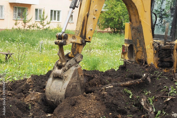 Obraz An excavator digs a hole in the ground. Summer, grass, dandelions, a wooden bench and an apartment building..