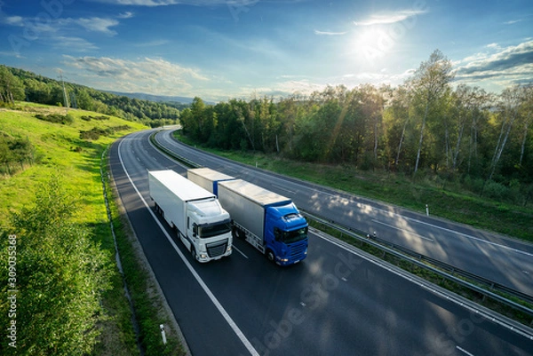 Fototapeta Niebieska ciężarówka wyprzedzająca białą ciężarówkę na asfaltowej autostradzie między lasami w promieniującym słońcu. Widok z góry.