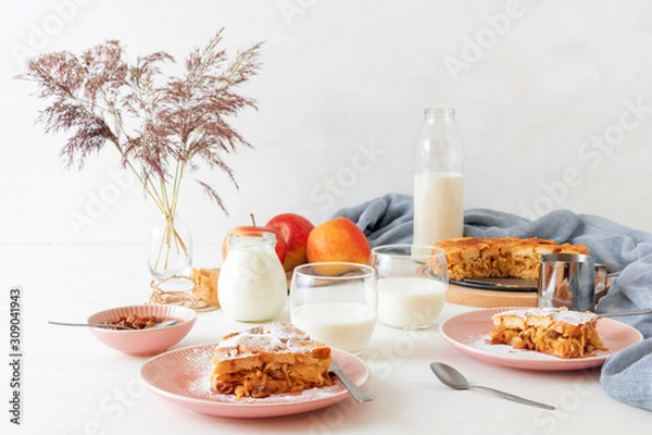 Fototapeta White wooden table with apple bread and butter pudding, jar with sour cream, glass of milk, background decorated with few apples and vase with dry grass.