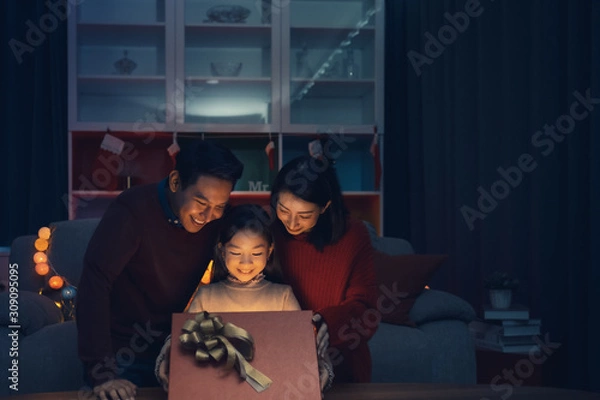 Fototapeta Happy family and little girl open magic gift box with light shining in living room that decorated with Christmas tree for Christmas festival coming soon, Asian Christmas family concept