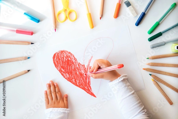 Fototapeta Close up of child's hands drawing red heart at white paper within colorful pens and pencils.