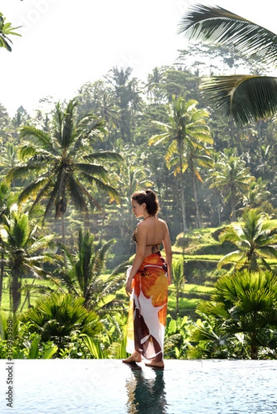 Obraz A young girl in an orange bathing suit stands on the edge of the pool against the backdrop of tropical greenery. Bali Indonesia. Ubud.