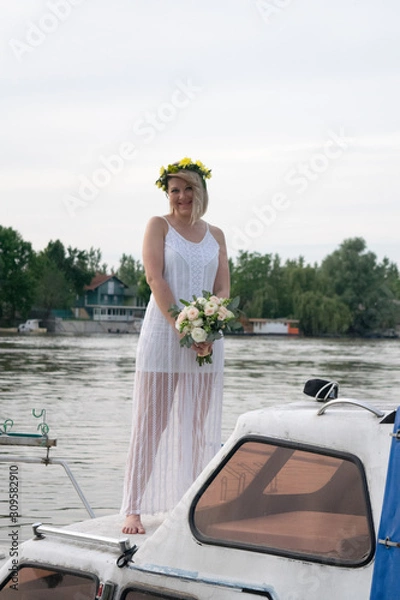 Fototapeta beautiful bride with wedding bouquet standing on a boat and posing for the photographer near the river