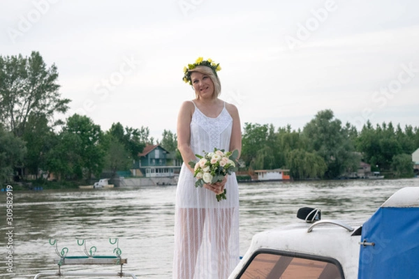 Fototapeta beautiful bride with wedding bouquet standing on a boat and posing for the photographer near the river