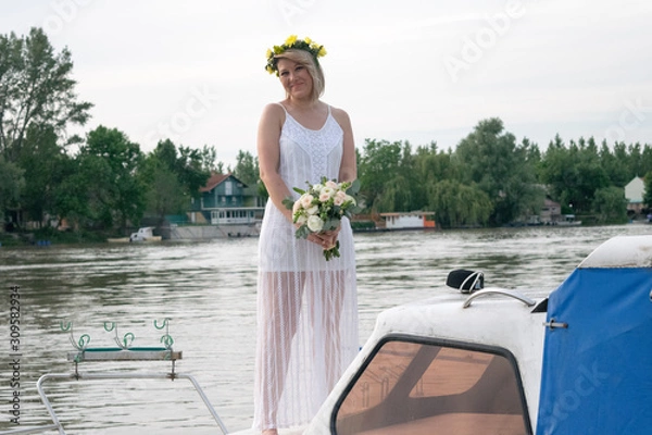 Fototapeta beautiful bride with wedding bouquet standing on a boat and posing for the photographer near the river