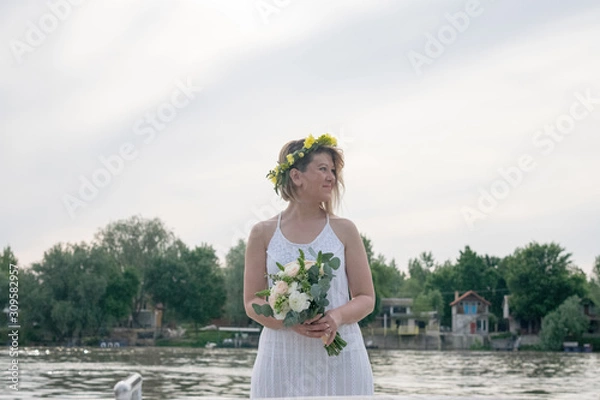 Fototapeta beautiful bride with wedding bouquet standing on a boat and posing for the photographer near the river
