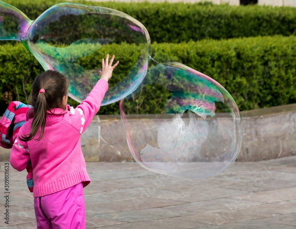 Obraz A little girl in pink is trying to reach and burst a large soap bubble. The view from the back.