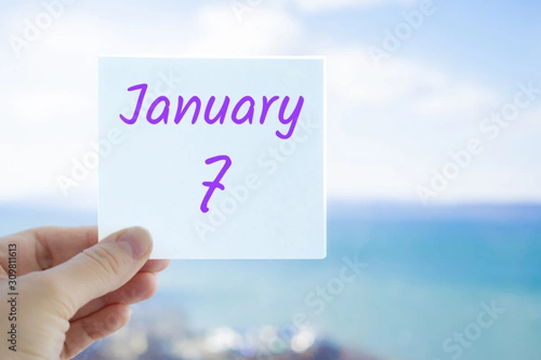 Obraz January 7th. Christmas day. Hand holding sticker with text January 7 on the blurred background of the sea and sky. Copy space for text. Month in calendar concept