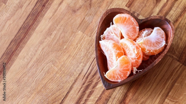 Fototapeta tangerine slices in a wooden bowl in the form of a heart on a wooden background top view. tangerines are flat lying. mandarins and place for text.
