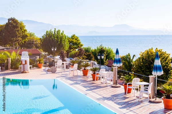 Fototapeta Outdoor pool with vibrant crystal water, parasols and deck chairs located on the coast of Garda with lake, hills and sun on background