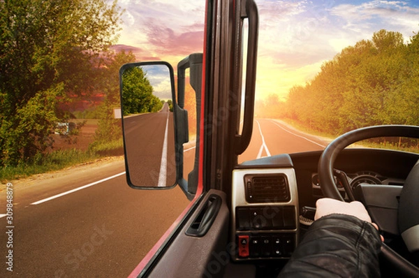 Fototapeta Truck dashboard with driver's hand on the steering wheel and side rear-view mirror on the countryside road against sky with sunset