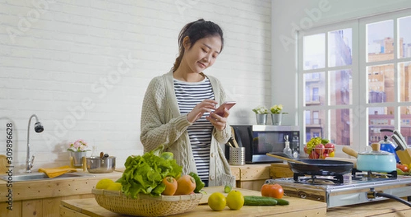 Obraz Picture of beautiful asian chinese woman cooking vegetables in cozy wooden kitchen while looking recipe on smartphone. young girl browsing website on tutor cooking class and smiling checking fruits.