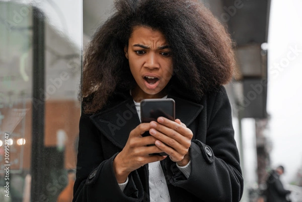 Fototapeta Oh my god, a girl looks in surprise at the phone with a shocked expression on her face, a reaction to a message or news about a cash flow to a bank account
