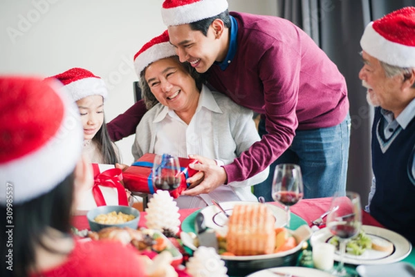 Fototapeta Big Asian family celebration in Christmas day, man hold gift box on hand and hug mother and which smiling and felling happy at home after breakfast time. Merry Xmas and happy new year.