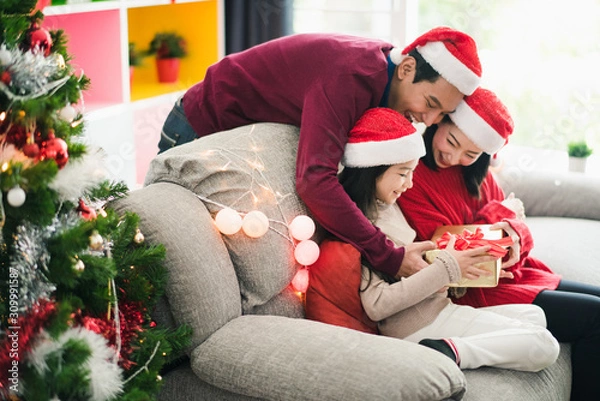 Obraz Young Asian family celebration in Christmas day, mother and daughter siting on sofa which hold give box, father stand back side,smiling felling happy in living room at home. Merry Xmas happy new year.