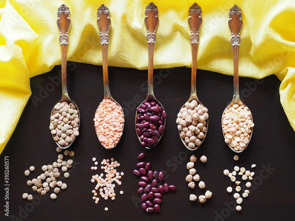 Fototapeta Beans, lentils, yellow peas, chickpeas in gold tablespoons with a yellow napkin on a black background closeup. Colorful picture with vegetarian legumes for decoration and design, top view.