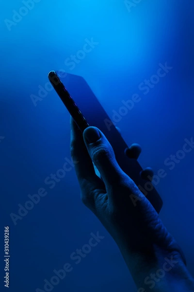 Fototapeta Hand holds smartphone. Neon classic blue light, vertical photo. Copyspace. Technology concept, future, social networks, content, mobile.