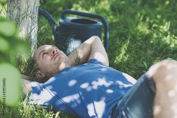 Fototapeta Man in blue t-shirt lying on grass with hands behind head in the garden with watering can nearby on greenery background on a sunny summer day. Free time and shying away from work concept. Toned